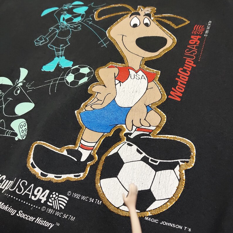 May include: Black t-shirt with a cartoon dog wearing a red, white, and blue soccer jersey and kicking a soccer ball. The text "World Cup USA94" is printed on the shirt.