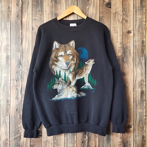 May include: Black sweatshirt with a wolf-themed graphic. The design includes three wolves, a full moon, and evergreen trees. The wolves are in shades of brown, grey, and white. The sweatshirt is displayed on a wooden hanger against a wooden backdrop.
