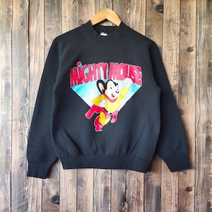 Op de afbeelding: Zwart sweatshirt met het stripfiguur Mighty Mouse in een ruit met een blauwe en gele achtergrond. De woorden "MIGHTY MOUSE" zijn in rood en wit. Het sweatshirt hangt aan een houten hanger.