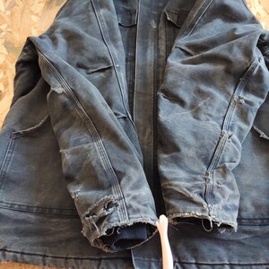 K&ouml;nnte beinhalten: Abgenutzte, dunkelblaue Arbeitsjacke mit sichtbaren Gebrauchsspuren. Die Jacke hat mehrere Taschen, eine Knopfleiste und abgenutzte Details an den &Auml;rmeln. Die Manschetten sind gerippt, und die Jacke scheint aus einem strapazierf&auml;higen Stoff zu sein.