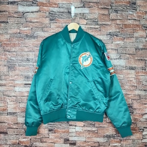 Puede incluir: Una chaqueta bomber de satén azul turquesa con un logotipo de delfín y las letras "M" y "NFL" bordadas en la parte delantera. La chaqueta tiene un cierre de presión y dos bolsillos.