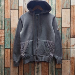 May include: A dark gray zip-up hoodie featuring a hood with matching drawstrings, a full-length front zipper, and two front pockets with quilted fabric panels. A small circular logo patch is visible on the upper left sleeve of the garment.