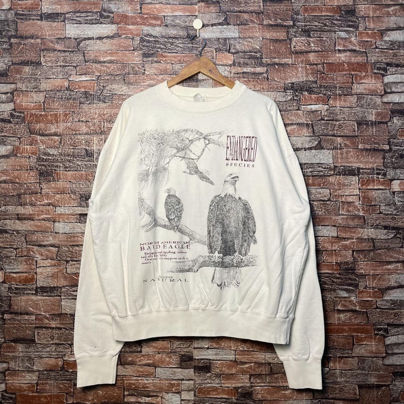 May include: White crewneck sweatshirt with a graphic print of two bald eagles in a forest setting. The text "Endangered Species" and "North American Bald Eagle" is printed on the sweatshirt.