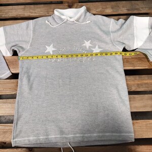 May include: Gray sweatshirt with a white collar and white trim on the sleeves. The sweatshirt has a white embroidered logo that says "Courage" with stars and the text "Paris".
