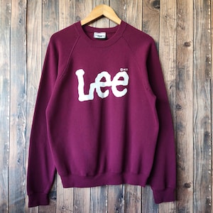 May include: A burgundy sweatshirt with the white word "Lee" across the chest. The long-sleeved top is hanging on a wooden hanger against a wooden plank background. The Lee logo is in a bold, blocky font.