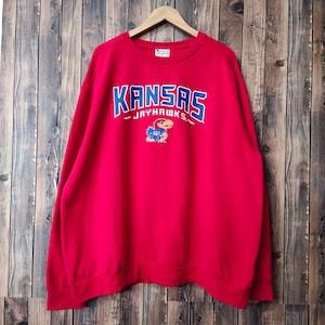 May include: Red sweatshirt with a blue and white Kansas Jayhawks logo. The logo features the words "Kansas" and "Jayhawks" with a cartoon Jayhawk mascot.