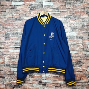 Könnte beinhalten: Eine marineblaue Collegejacke mit gelben und weißen Streifen an Kragen und Ärmelbündchen. Die Jacke hat einen Knopfverschluss und ein gesticktes Logo eines Maskottchens mit dem Wort "NAVY" darunter.