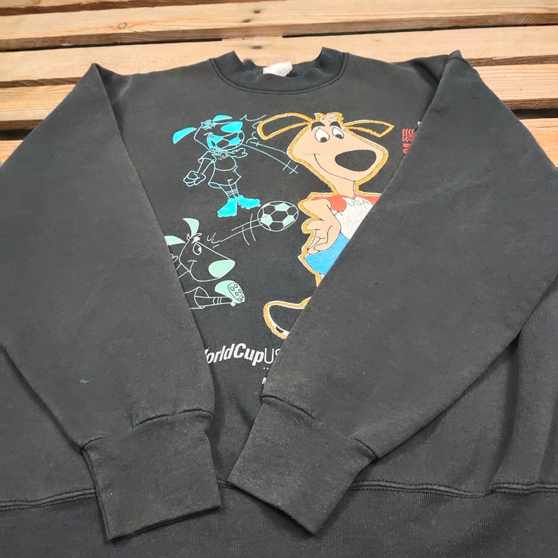 May include: Black sweatshirt with a graphic print of cartoon dogs playing soccer. The text "World Cup USA 94" is printed on the bottom of the sweatshirt.