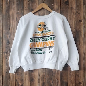 Puede incluir: Sudadera blanca con un logotipo verde y amarillo de los Edmonton Eskimos Grey Cup 87 Champions. El texto dice "Edmonton Eskimos Grey Cup '87 Champions" con la fecha "29 de noviembre de 1987" y la ubicación "B.C. Place Dome Stadium Vancouver". El marcador se indica como "Eskimos 38, Argonauts 36".