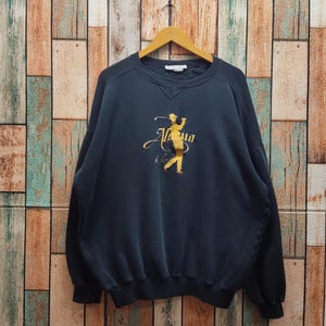 May include: Navy blue sweatshirt with a gold graphic of a golfer in action and the word "Aloha". The sweatshirt is hanging on a wooden hanger against a backdrop of vertical wooden planks.