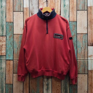 May include: A red long-sleeve pullover sweatshirt featuring a dark blue mock neck collar with a quarter-zip closure. A rectangular patch on the left chest displays the text CHAPS RALPH LAUREN 78, and a small black tag is on the left sleeve.