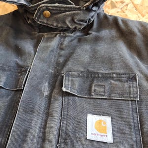 K&ouml;nnte beinhalten: Dunkelgraue Carhartt-Jacke mit Kapuze und mehreren Taschen. Die Jacke weist sichtbare Gebrauchsspuren auf. Das Carhartt-Logo befindet sich auf einem quadratischen Aufn&auml;her auf der Vordertasche. Hergestellt aus strapazierf&auml;higem, robustem Stoff.