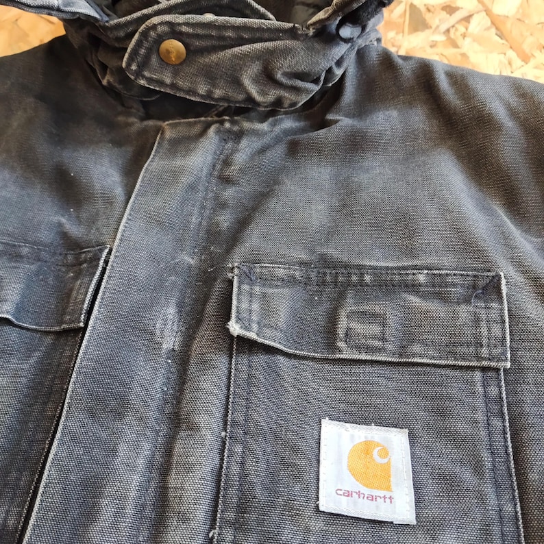 K&ouml;nnte beinhalten: Dunkelgraue Carhartt-Jacke mit Kapuze und mehreren Taschen. Die Jacke weist sichtbare Gebrauchsspuren auf. Das Carhartt-Logo befindet sich auf einem quadratischen Aufn&auml;her auf der Vordertasche. Hergestellt aus strapazierf&auml;higem, robustem Stoff.