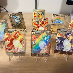 May include: A collection of gold-colored Pokémon trading cards, each displayed on a clear plastic stand. The cards feature various Pokémon characters, including Charizard, Pikachu, and Blastoise, with vibrant colors and detailed artwork. The cards have text and numbers.
