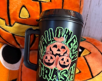 May include: Black travel mug with a handle, featuring a Halloween-themed design. The design includes three jack-o'-lanterns and the words "Halloween Trash" in green and orange. The mug is set against a backdrop of orange pumpkin plushies.