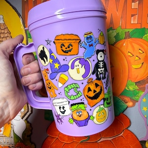 以下が含まれることがあります： 蓋とハンドルが付いた、紫色のハロウィーンをテーマにした大きなマグカップ。マグカップは、幽霊、カボチャ、魔女の帽子など、さまざまな漫画のハロウィーンのキャラクターで飾られています。マグカップは人の手で持たれています。