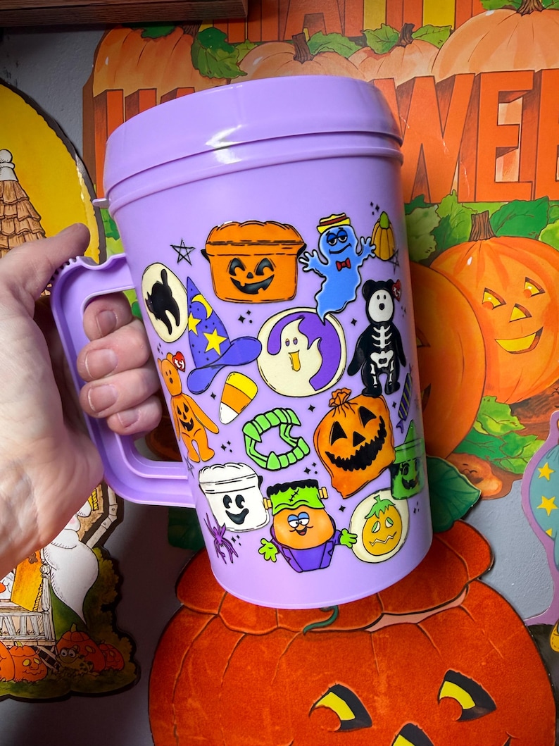 以下が含まれることがあります： 蓋とハンドルが付いた、紫色のハロウィーンをテーマにした大きなマグカップ。マグカップは、幽霊、カボチャ、魔女の帽子など、さまざまな漫画のハロウィーンのキャラクターで飾られています。マグカップは人の手で持たれています。