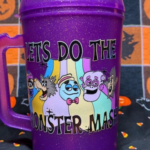 May include: A purple glittery plastic mug with a handle and a lid. The front features a colorful graphic of cartoon monsters and the text "LETS DO THE MONSTER MASH" in black lettering. The mug has a translucent finish with embedded gold glitter.