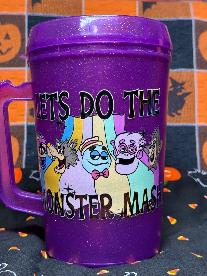 May include: A purple glittery plastic mug with a handle and a lid. The front features a colorful graphic of cartoon monsters and the text "LETS DO THE MONSTER MASH" in black lettering. The mug has a translucent finish with embedded gold glitter.