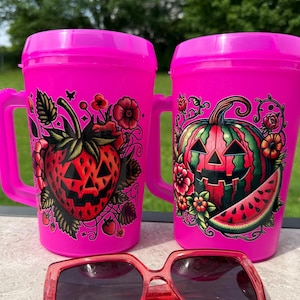 May include: Two bright pink plastic mugs with handles, each featuring a colorful graphic. One mug displays a jack-o'-lantern strawberry with flowers, while the other shows a jack-o'-lantern watermelon with flowers. Red-tinted sunglasses sit in front.