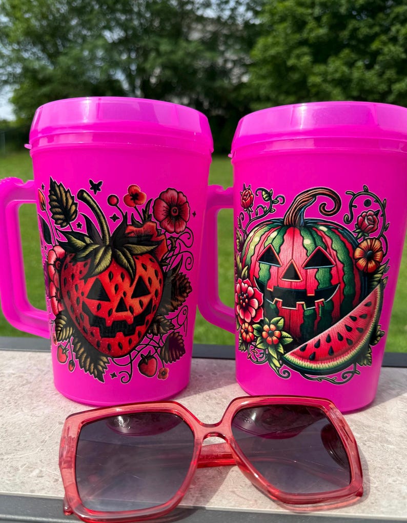May include: Two bright pink plastic mugs with handles, each featuring a colorful graphic. One mug displays a jack-o'-lantern strawberry with flowers, while the other shows a jack-o'-lantern watermelon with flowers. Red-tinted sunglasses sit in front.