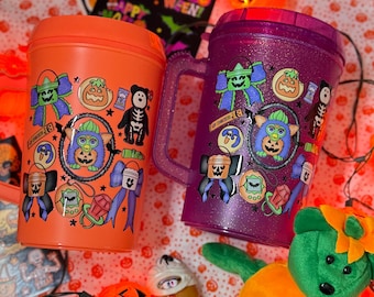 Peut inclure: Un gobelet en plastique orange et une tasse en plastique violette à paillettes, tous deux ornés d'autocollants colorés sur le thème d'Halloween représentant un Furby, un ours squelette, des citrouilles et des nœuds, avec un ours en peluche vert.
