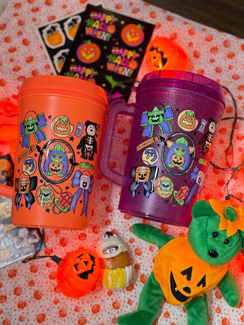 Puede incluir: Un vaso naranja y un vaso morado con purpurina, ambos con pegatinas de Halloween de un Furby, lazos, calabazas y esqueletos. El vaso naranja tiene tapa, mientras que el morado tiene asa. Se incluye un oso de peluche verde con disfraz.