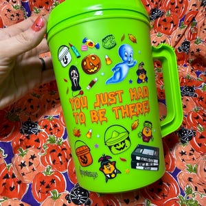 Op de afbeelding: Een fel limoengroene plastic beker met een handvat, voorzien van diverse kleurrijke Halloween-stickers waaronder een geest, een pompoen en snoepjes. De tekst "YOU JUST HAD TO BE THERE!" is in het oranje gedrukt, met "Goosebumps" onderaan.