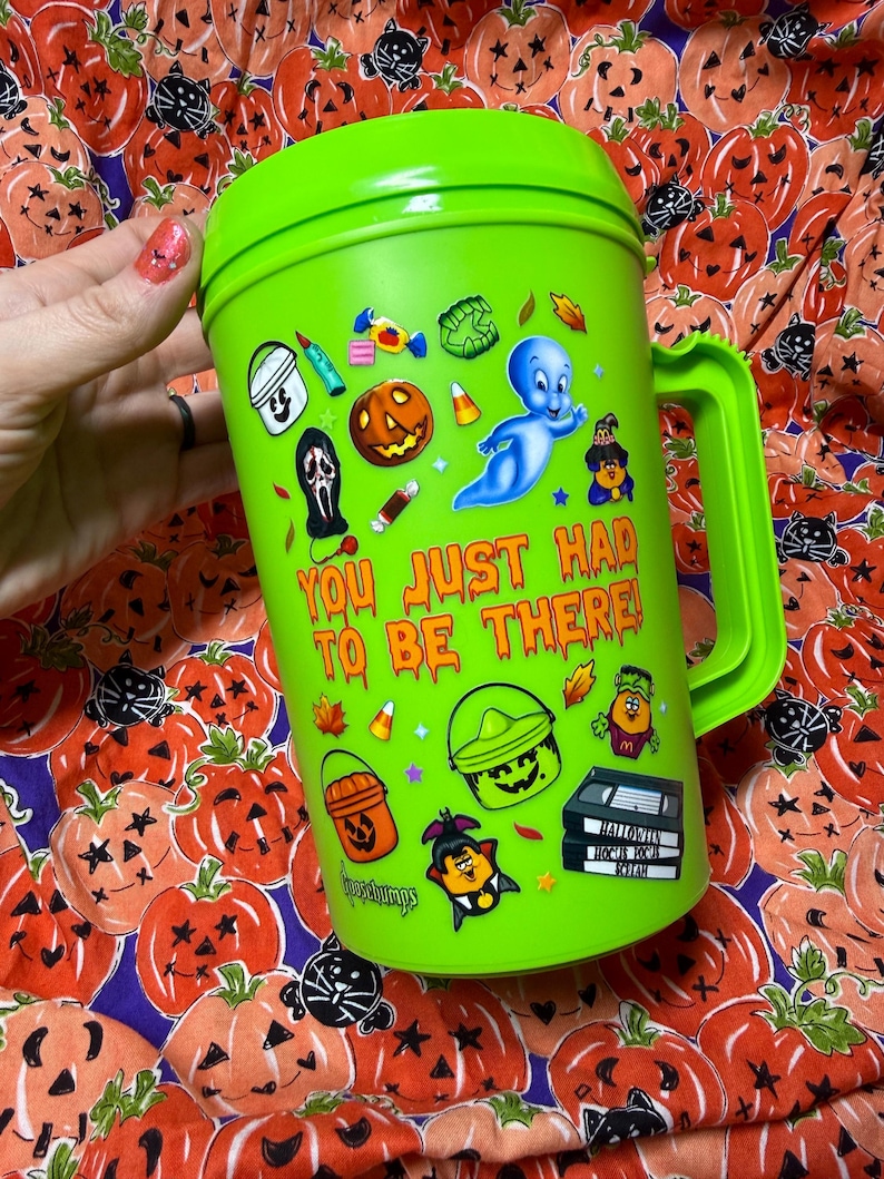 Op de afbeelding: Een fel limoengroene plastic beker met een handvat, voorzien van diverse kleurrijke Halloween-stickers waaronder een geest, een pompoen en snoepjes. De tekst "YOU JUST HAD TO BE THERE!" is in het oranje gedrukt, met "Goosebumps" onderaan.
