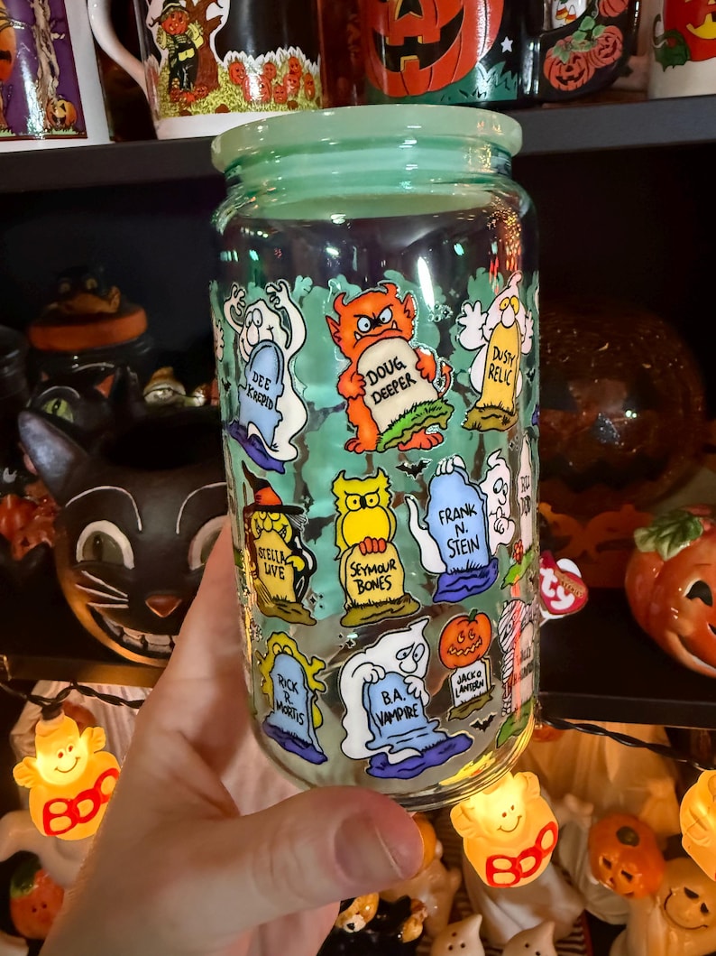 Peut inclure: Un bocal en verre transparent avec un couvercle vert clair, d&eacute;cor&eacute; de personnages de dessins anim&eacute;s d'Halloween sur des pierres tombales. Le bocal pr&eacute;sente des personnages comme "Doug Deeper" et "B.A. Vampire".