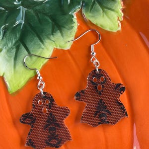 Peut inclure: Une paire de boucles d'oreilles sur le thème d'Halloween. Chaque boucle d'oreille présente un charme en forme de fantôme avec un motif de dentelle noire sur un fond orange. Les boucles d'oreilles ont des crochets d'oreille argentés.