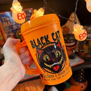 May include: An orange plastic mug with a lid and handle featuring a graphic of a black cat face with green eyes and the text "BLACK CAT CAULDRON & BROOM POLISH BE READY FOR THE WITCHING HOUR" printed in black on the front of the container.