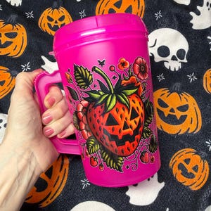 May include: A bright pink plastic travel mug with a handle and a lid. The mug features a graphic illustration of a red strawberry designed with a jack-o'-lantern face, surrounded by green leaves, small red flowers, and tiny star accents.