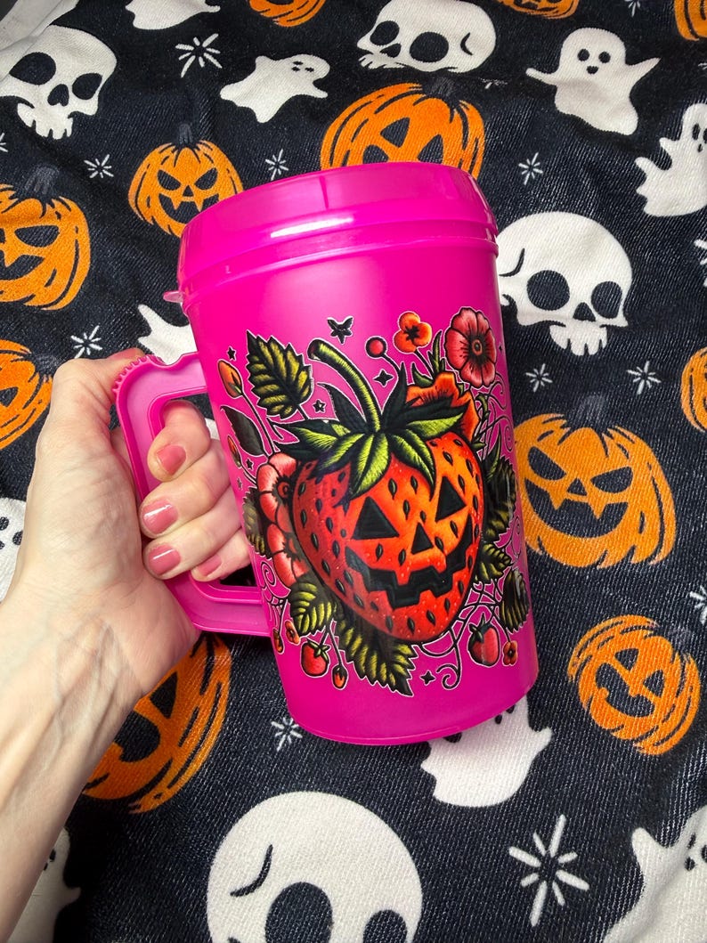May include: A bright pink plastic travel mug with a handle and a lid. The mug features a graphic illustration of a red strawberry designed with a jack-o'-lantern face, surrounded by green leaves, small red flowers, and tiny star accents.