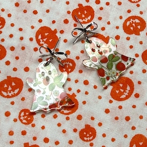 Peut inclure: Une paire de boucles d'oreilles en forme de fantôme avec une base en acrylique transparent ornée d'un motif imprimé de fraises rouges, de feuilles vertes et de fleurs blanches. Chaque boucle est surmontée d'un petit nœud en métal argenté.