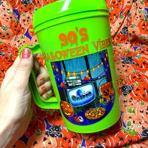 Op de afbeelding: Een fel limoengroene plastic beker met een handvat en deksel. De beker is voorzien van de tekst 90's HALLOWEEN VIBES in oranje en een grafische illustratie van een televisiescherm met het woord CASPER, een pompoen, een pizza en lichtsnoeren.
