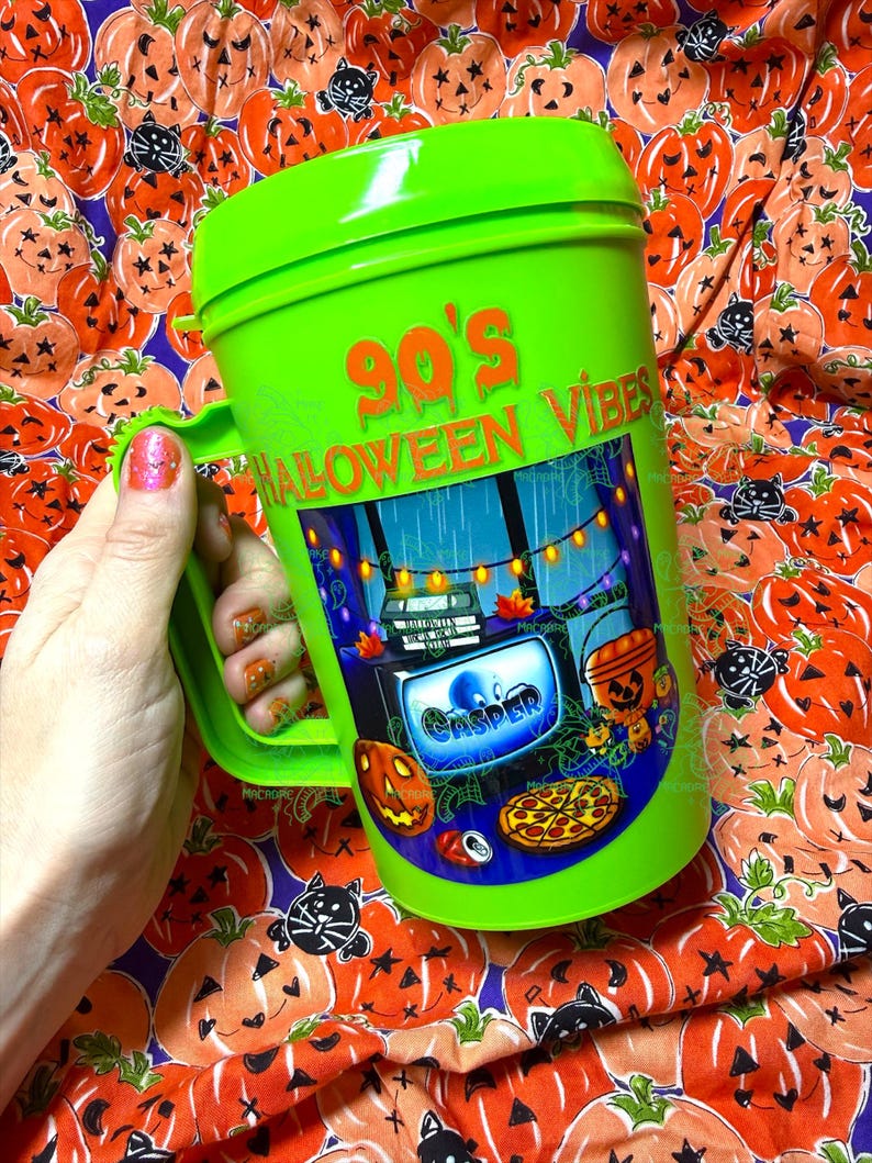 Op de afbeelding: Een fel limoengroene plastic beker met een handvat en deksel. De beker is voorzien van de tekst 90's HALLOWEEN VIBES in oranje en een grafische illustratie van een televisiescherm met het woord CASPER, een pompoen, een pizza en lichtsnoeren.