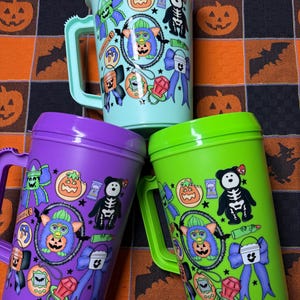 May include: Three plastic mugs in light blue, purple, and lime green, each featuring a repeating graphic print of a Furby, a skeleton bear, a jack-o'-lantern, a bow, a ring, and a digital pet toy, all with matching lids and side handles.