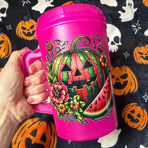 May include: A bright pink plastic mug with a handle and lid, featuring a colorful graphic of a jack-o'-lantern carved from a watermelon, surrounded by red and orange flowers, green leaves, and a slice of watermelon on the front surface.
