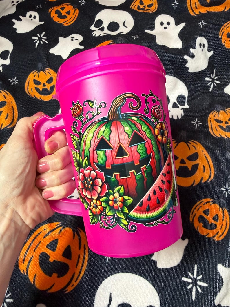 May include: A bright pink plastic mug with a handle and lid, featuring a colorful graphic of a jack-o'-lantern carved from a watermelon, surrounded by red and orange flowers, green leaves, and a slice of watermelon on the front surface.