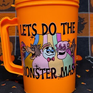 May include: A bright orange plastic mug with a handle and a lid. The front features a colorful graphic of five cartoon monsters and the black text LETS DO THE MONSTER MASH. The design includes a mummy, a werewolf, a ghost, and other creatures.