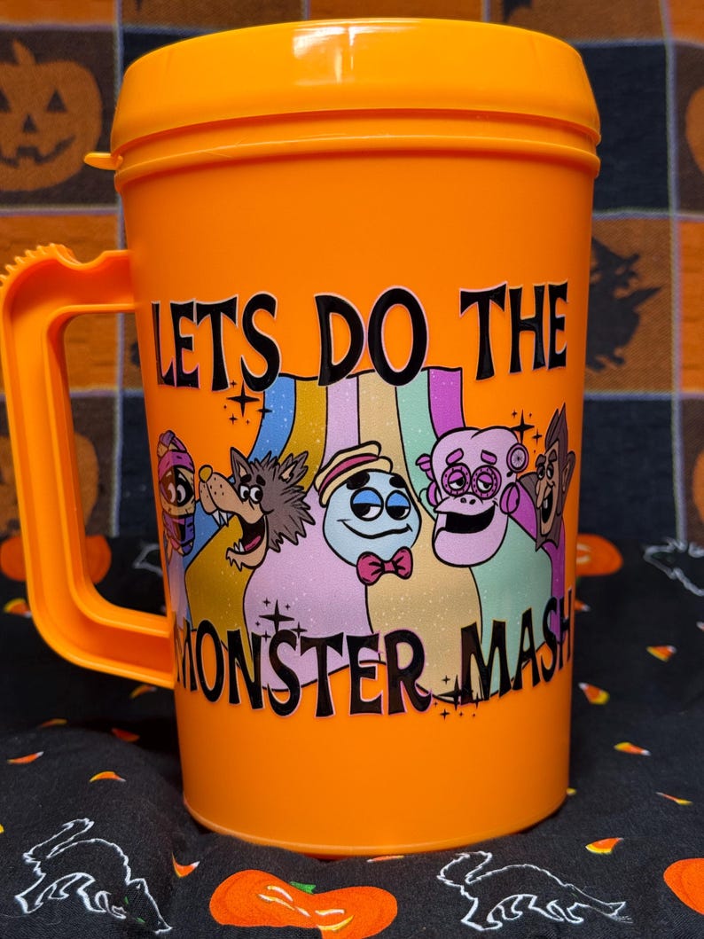 May include: A bright orange plastic mug with a handle and a lid. The front features a colorful graphic of five cartoon monsters and the black text LETS DO THE MONSTER MASH. The design includes a mummy, a werewolf, a ghost, and other creatures.