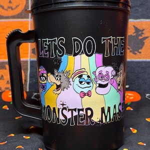 May include: A black plastic travel mug with a handle and lid, featuring a colorful graphic of five cartoon monsters and the text "LETS DO THE MONSTER MASH" printed in a bold, stylized font across the front of the container.