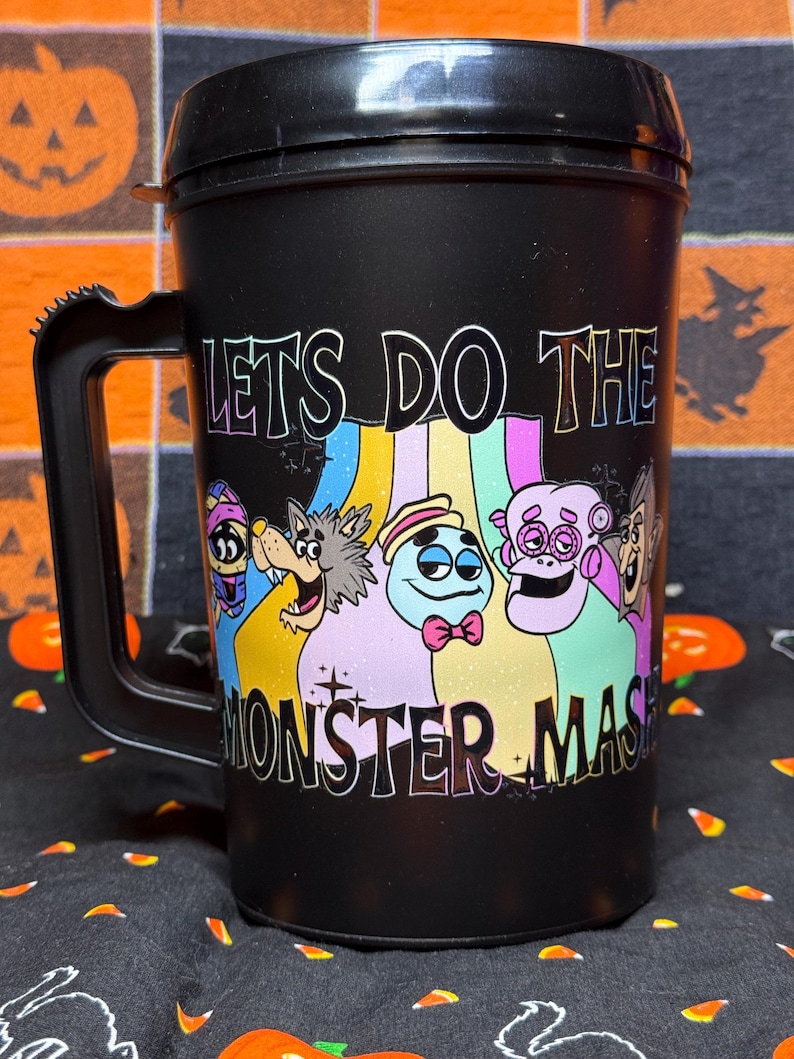 May include: A black plastic travel mug with a handle and lid, featuring a colorful graphic of five cartoon monsters and the text "LETS DO THE MONSTER MASH" printed in a bold, stylized font across the front of the container.