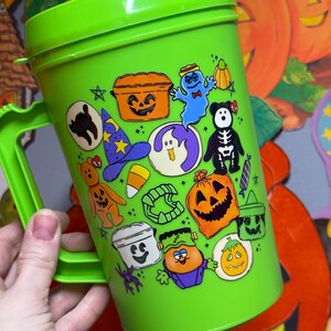 以下が含まれることがあります： 明るい緑色のハロウィーンをテーマにしたプラスチックカップ、ハンドル付き。カップは、カボチャ、幽霊、黒猫など、さまざまなハロウィーンのキャラクターの漫画の画像で飾られています。カップには蓋が付いています。