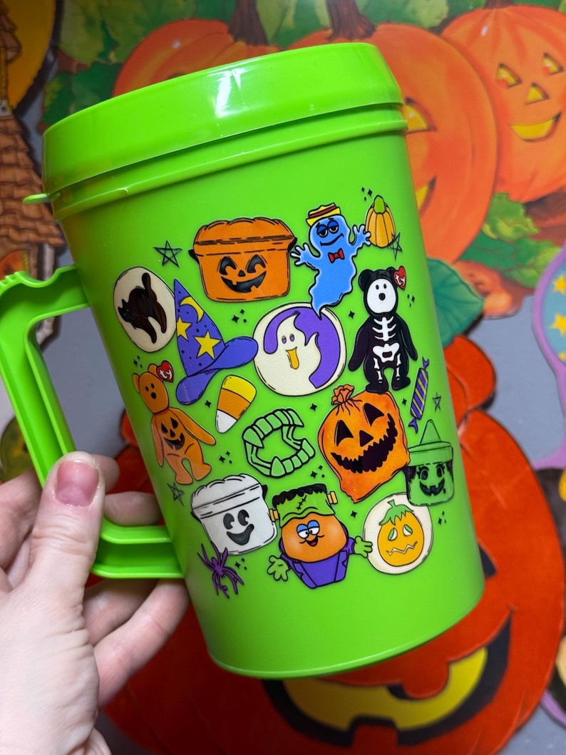 以下が含まれることがあります： 明るい緑色のハロウィーンをテーマにしたプラスチックカップ、ハンドル付き。カップは、カボチャ、幽霊、黒猫など、さまざまなハロウィーンのキャラクターの漫画の画像で飾られています。カップには蓋が付いています。