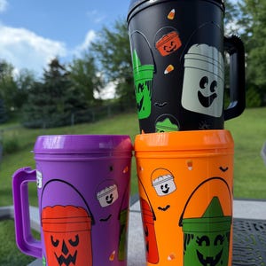 May include: Three plastic travel mugs with handles and lids, featuring a Halloween theme. One mug is black, one is purple, and one is orange. The mugs are decorated with colorful patterns of jack-o'-lantern buckets, candy corn, and small spiders.