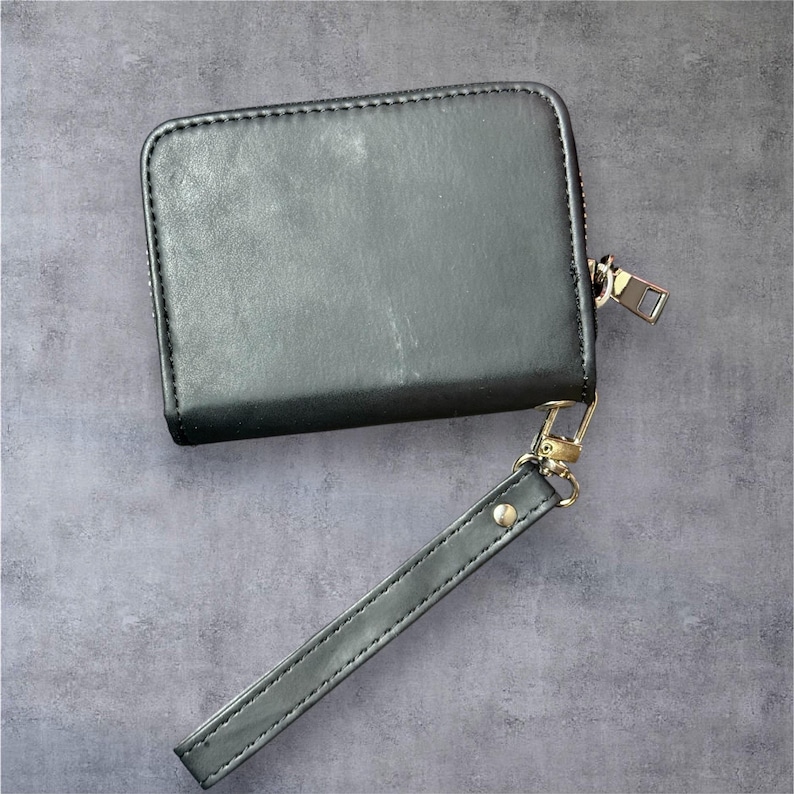 K&ouml;nnte beinhalten: Eine schwarze, quadratische Geldb&ouml;rse mit Rei&szlig;verschluss und Handschlaufe. Die Geldb&ouml;rse besteht aus einem glatten Material und hat einen goldfarbenen Rei&szlig;verschluss und Beschl&auml;ge. Die Handschlaufe ist mit einem goldfarbenen Verschluss befestigt.