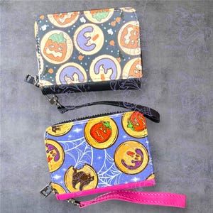 May include: Two Halloween-themed wallets. The top wallet has a dark background with orange pumpkin and purple ghost designs. The bottom wallet has a blue background with a spiderweb design and a pink wrist strap. Both wallets have a zipper closure.