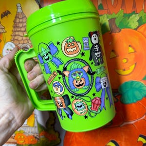 Puede incluir: Una taza de viaje de pl&aacute;stico de color verde lima brillante con asa y tapa, que presenta un estampado colorido alrededor con varios iconos tem&aacute;ticos de Halloween, incluyendo un personaje Furby central, calabazas y lazos.